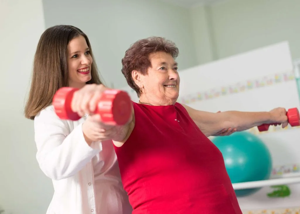 A physical therapist assists an elderly woman with a weightlifting exercise, using light dumbbells during a rehabilitation session